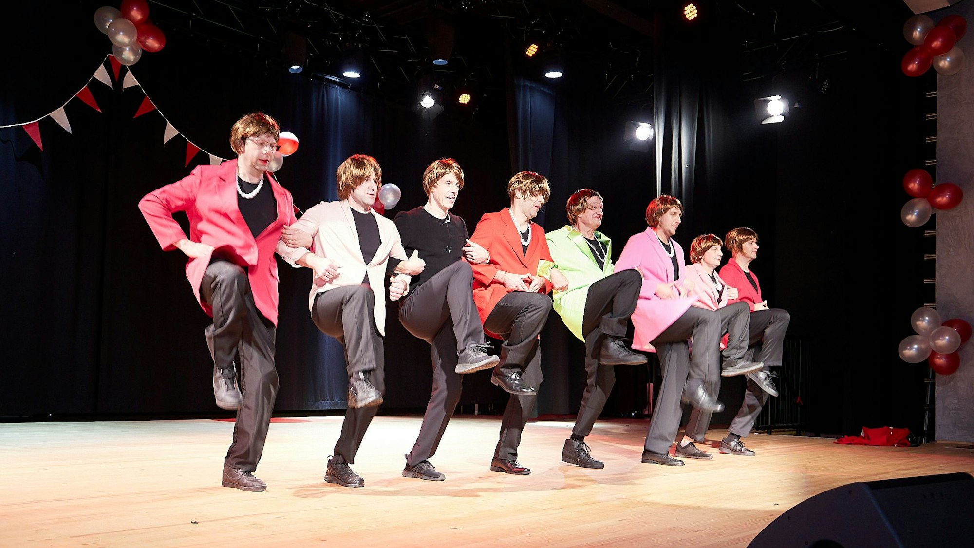 Acht Männer mit Angela-Merkel-Perrücken heben die Beine beim Tanzen auf einer Bühne an.