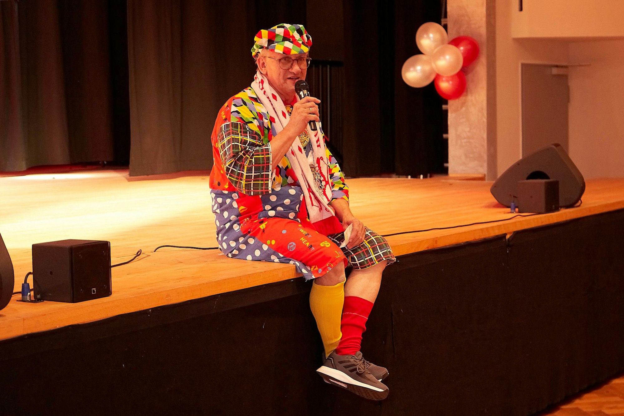 Ein älterer Herr im Karnevalskostüm mit unterschiedlichen Strümpfen sitzt am Bühnenrand und spricht in ein Mikrofon, das er hält.