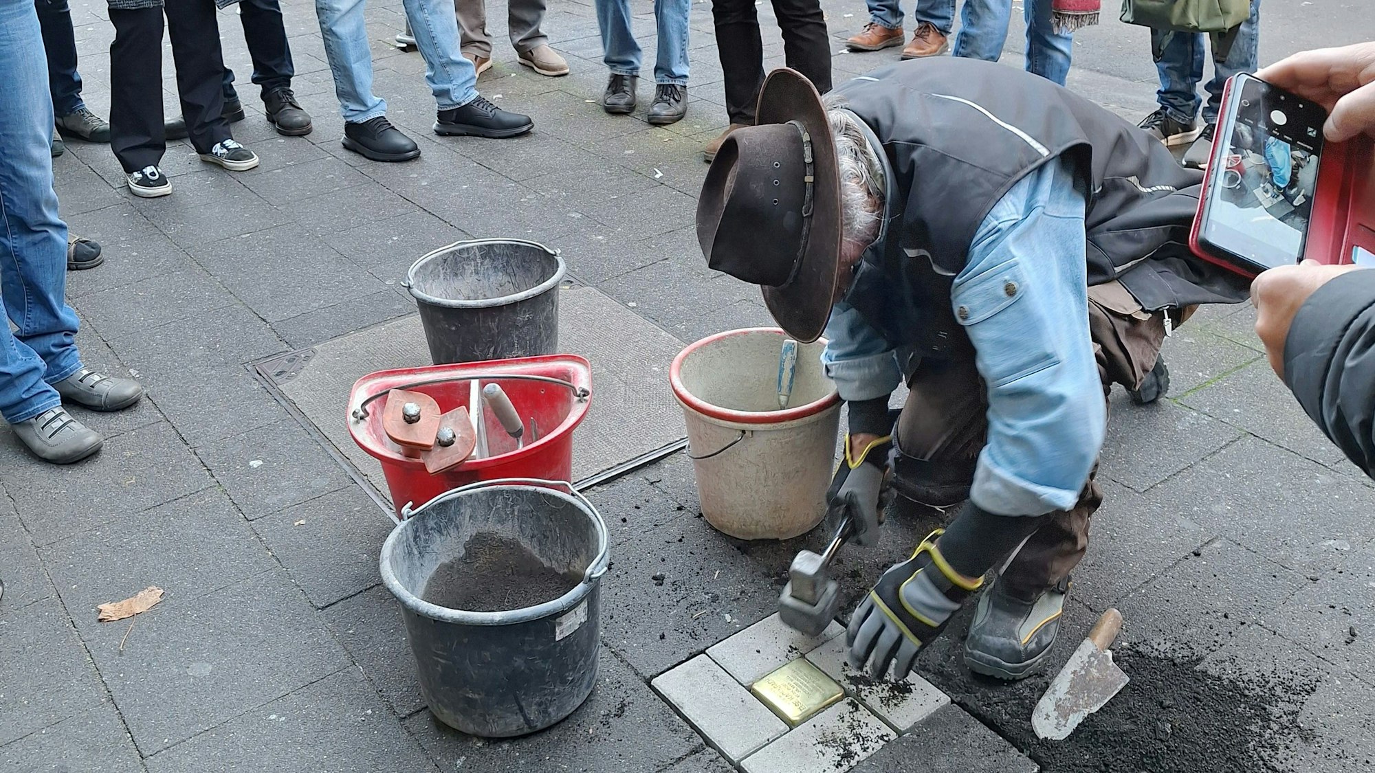 Ein Mann mit Hut arbeitet an einem Bürgersteig. Er ist dabei, einen glänzenden Erinnerungsstein zu verlegen.