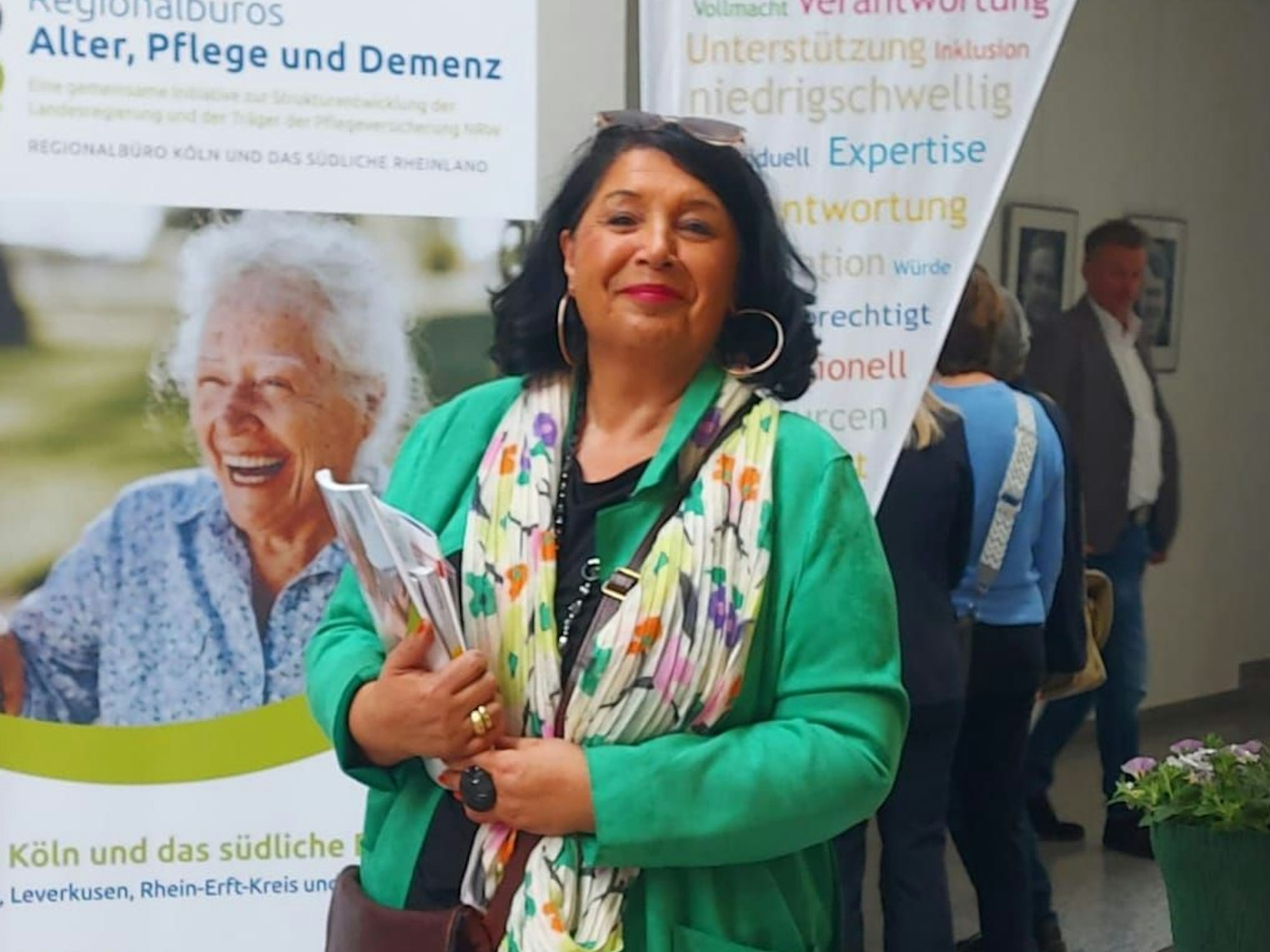 Fatma Dik-Thiel steht vor einer Werbewand der Regionalbüros „Alter, Pflege und Demenz“, in der Hand hält sie Broschüren.