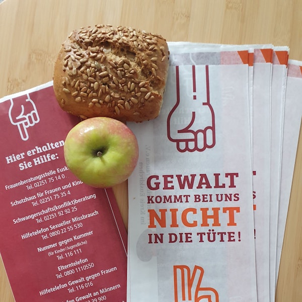 Mehrere Brötchentüten liegen auf einem Tisch, daneben ein Apfel und ein Körnerbrötchen. Auf der Vorderseite der Tüten steht „Gewalt kommt bei uns nicht in die Tüte“, auf der Rückseite sind Telefonnummern verschiedener Anlaufstellen, falls man von Gewalt betroffen ist.