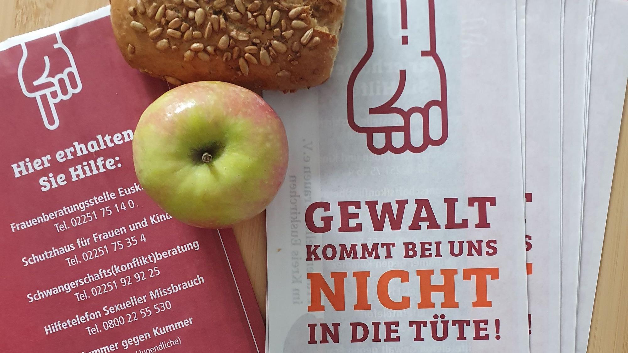 Mehrere Brötchentüten liegen auf einem Tisch, daneben ein Apfel und ein Körnerbrötchen. Auf der Vorderseite der Tüten steht „Gewalt kommt bei uns nicht in die Tüte“, auf der Rückseite sind Telefonnummern verschiedener Anlaufstellen, falls man von Gewalt betroffen ist.