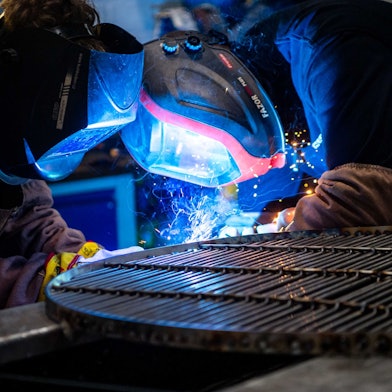 Zwei Jugendliche stehen mit Schutzmasken an einem Eisengitter und schweißen etwas. Ihre Gesichter sind in blaues Licht getaucht und es sprühen Funken.