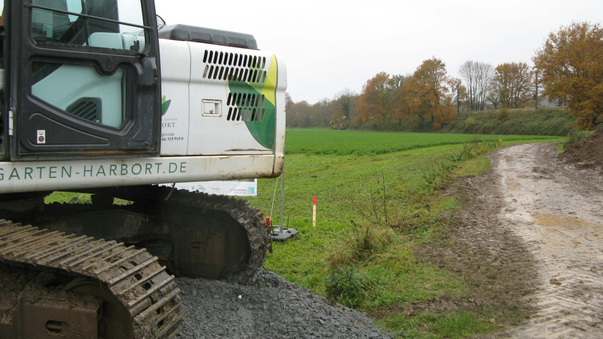 Ein matschiger Weg zwischen einem Feld und einem Bahndamm. Links im Vordergrund steht ein Bagger.