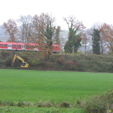 Ein Bagger steht vor einem Bahndamm, auf dem ein S-Bahnzug fährt. Ein Traktor zieht einen beladenen Anhänger weg.