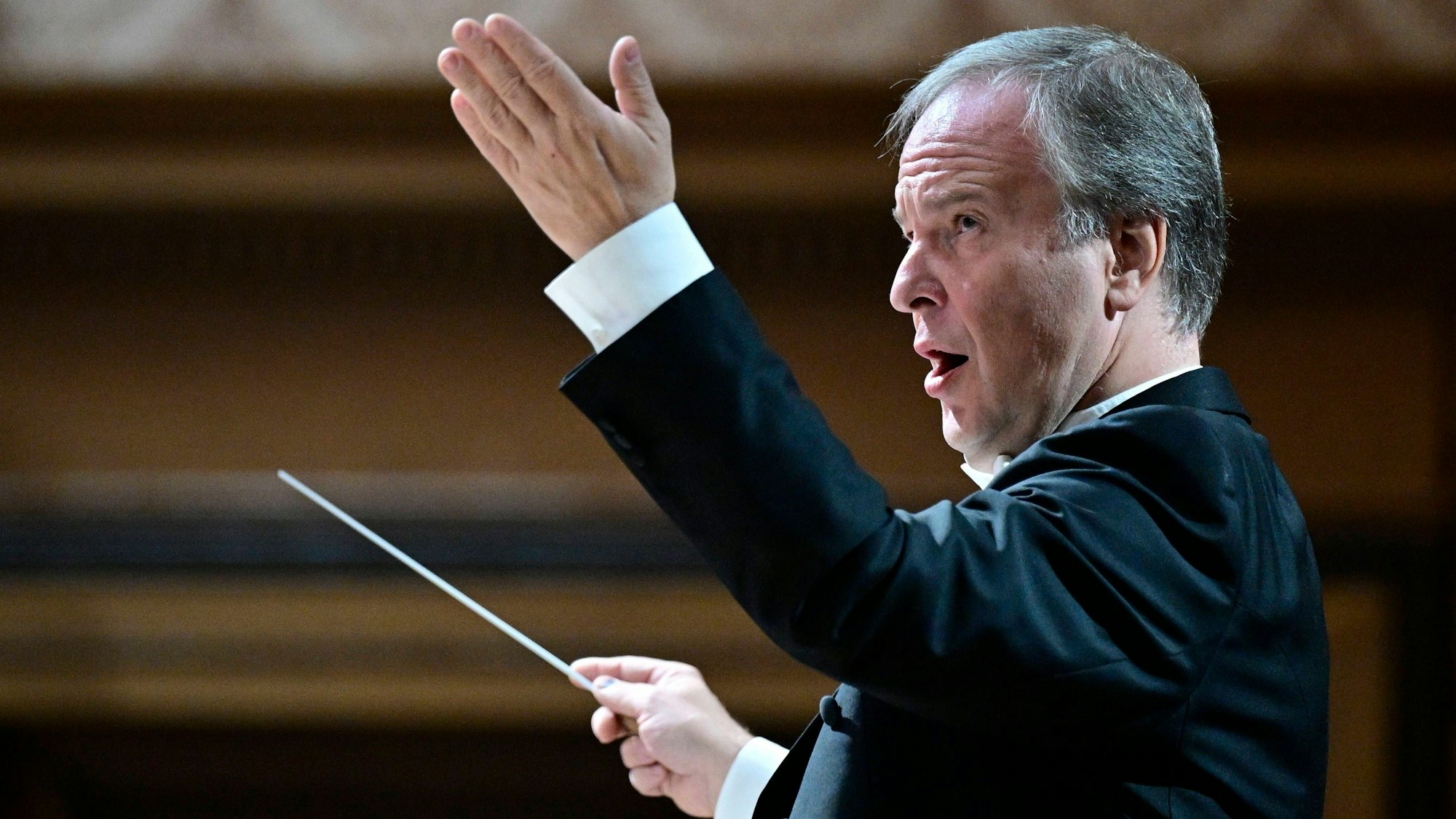 Ein Dirigent hebt mit forderndem Blick seine linke Hand und hält in der anderen einen Taktstock.