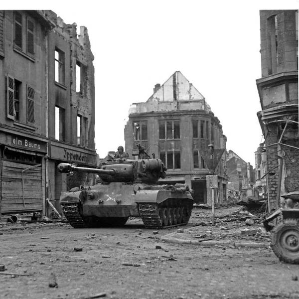 Eine historische Aufnahme zeigt einen Panzer, umgeben von zerstörten Häusern. Am Straßenrand steht ein Soldat.