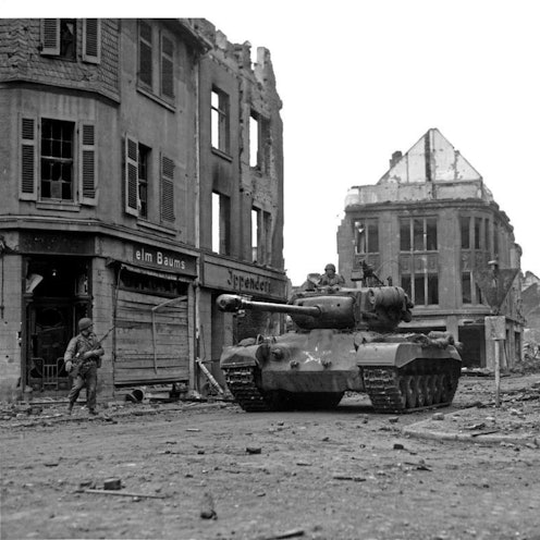 Eine historische Aufnahme zeigt einen Panzer, umgeben von zerstörten Häusern. Am Straßenrand steht ein Soldat.