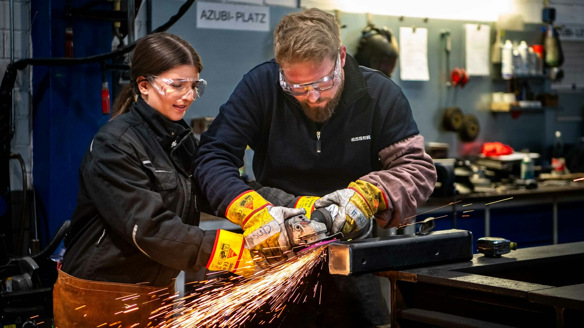 Eine Schülerin steht mit Schutzbrille an einer Werkbank und hält zusammen mit einem Mann eine Schleifmaschine an ein Metallstück. Es fliegen viele Funken.