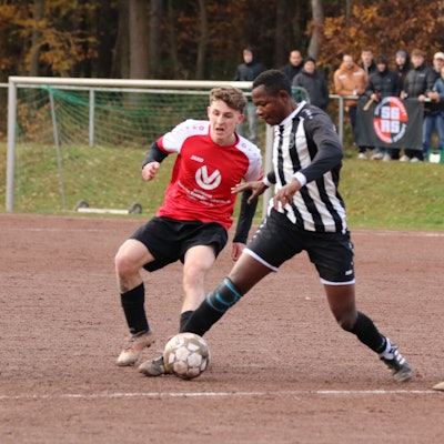 Zwei Fußballer im Zweikampf auf einem Aschenplatz. Im Hintergrund sind die Zuschauer zu erkennen.