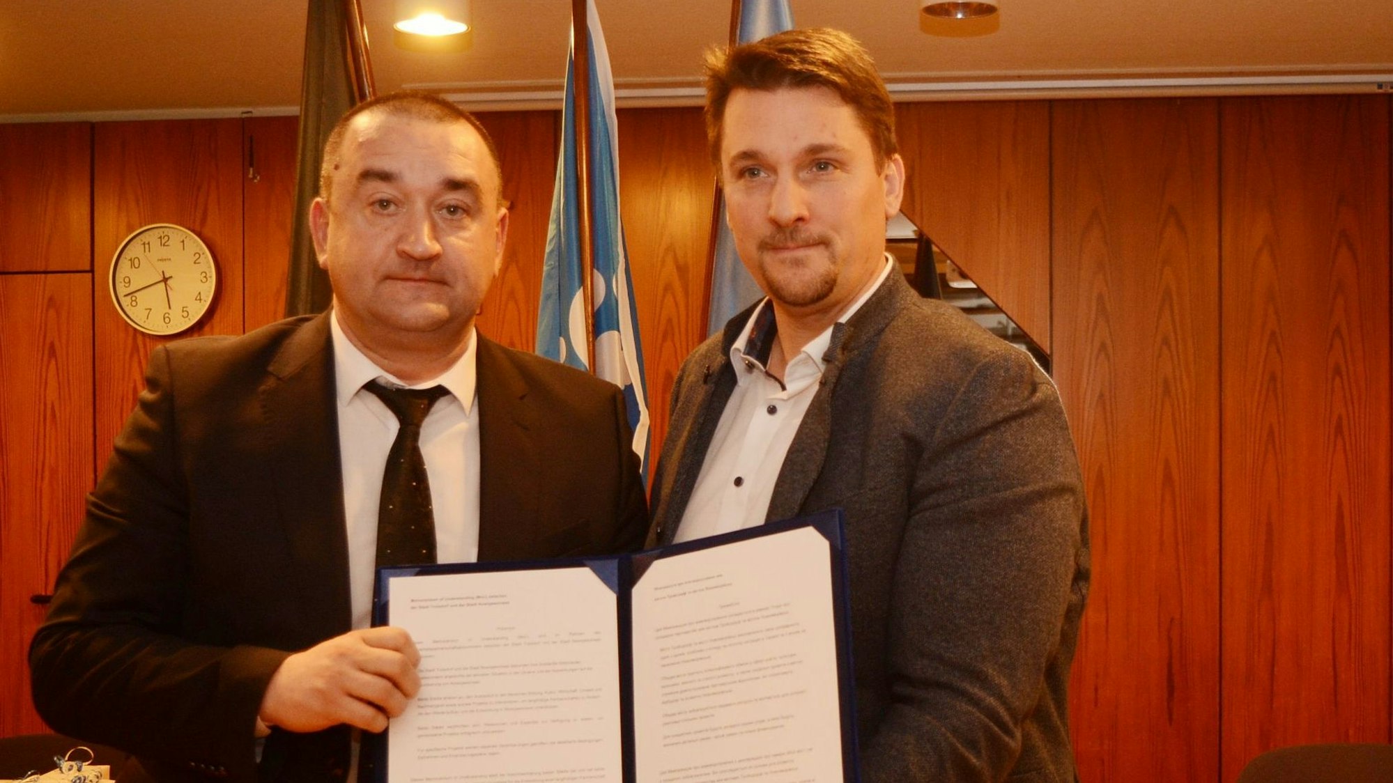 Zwei Männer im Anzug zeigen eine geöffnete Urkundenmappe.