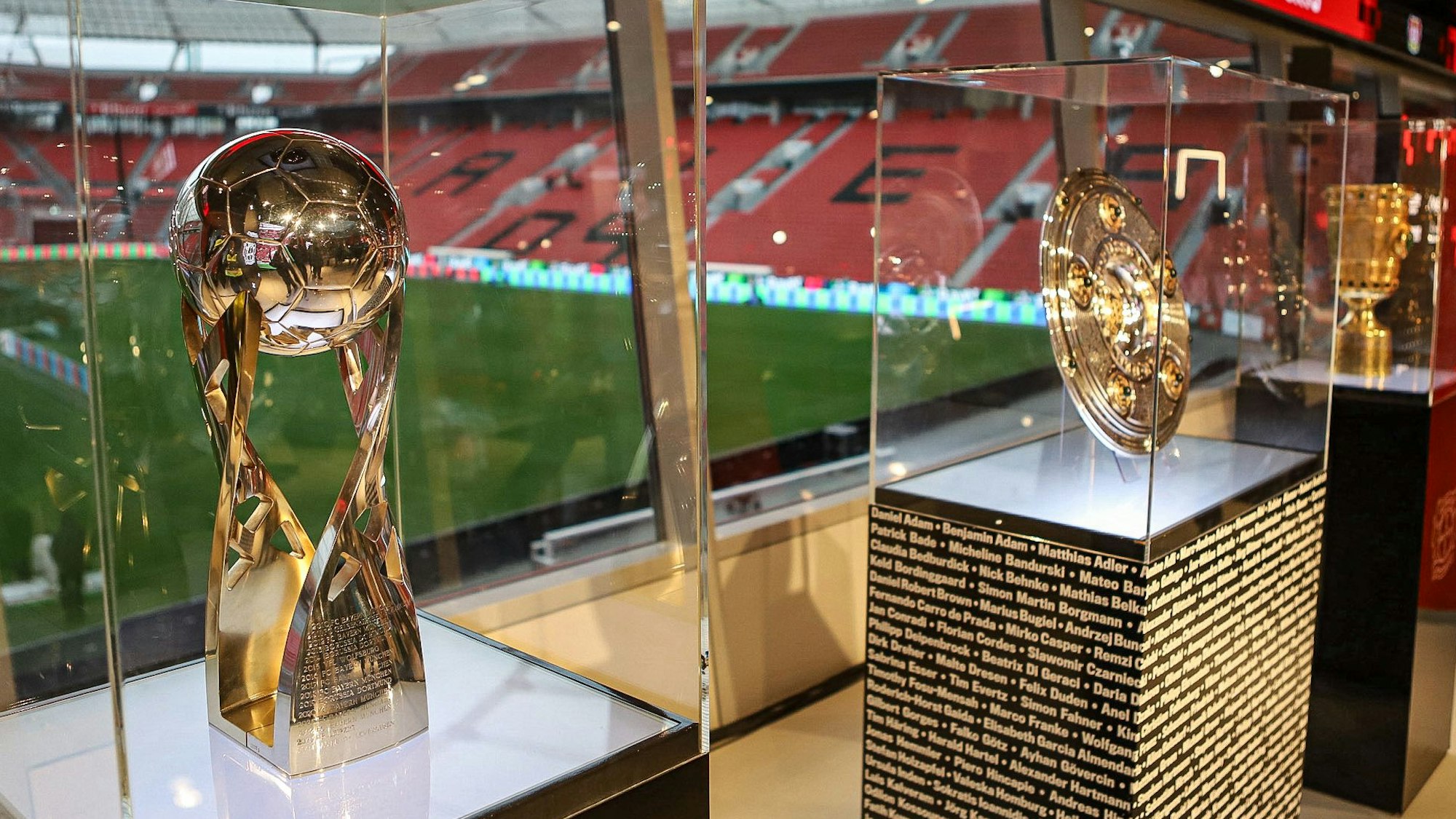 Die drei Pokale aus der vorigen Saison stehen vor einem Fenster in der Bay-Arena.