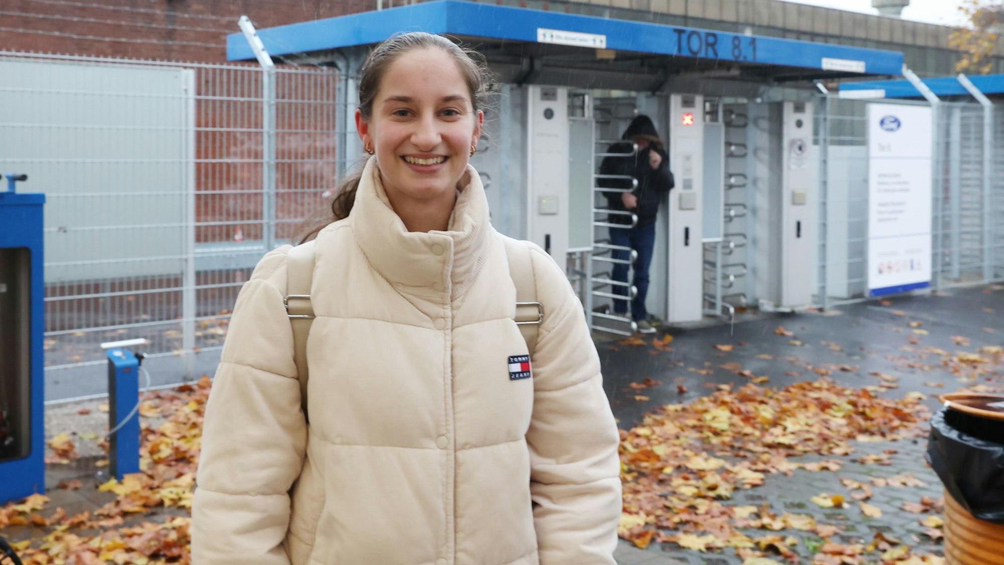 Sophia Braun steht in einer beigefarbenen Winterjacke vor einem Zaun mit Drehkreuz.