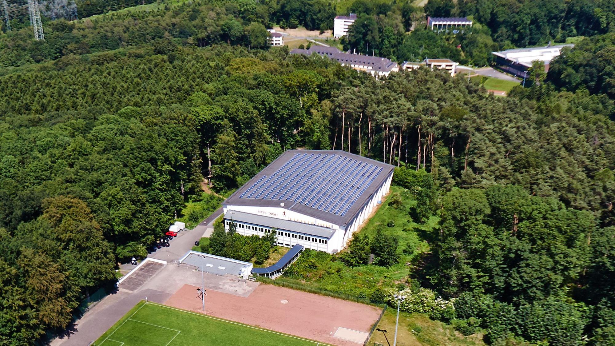 Blick aus der Vogelperspektive auf einen Fußballplatz, zwei Flachbauten und eine große Sporthalle, die von Wald umgeben ist.