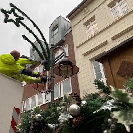 Zu sehen ist, wie zwei Stadtwerke-Mitarbeiter die Weihnachtsbeleuchtung an einem Laternenmast anbringen.