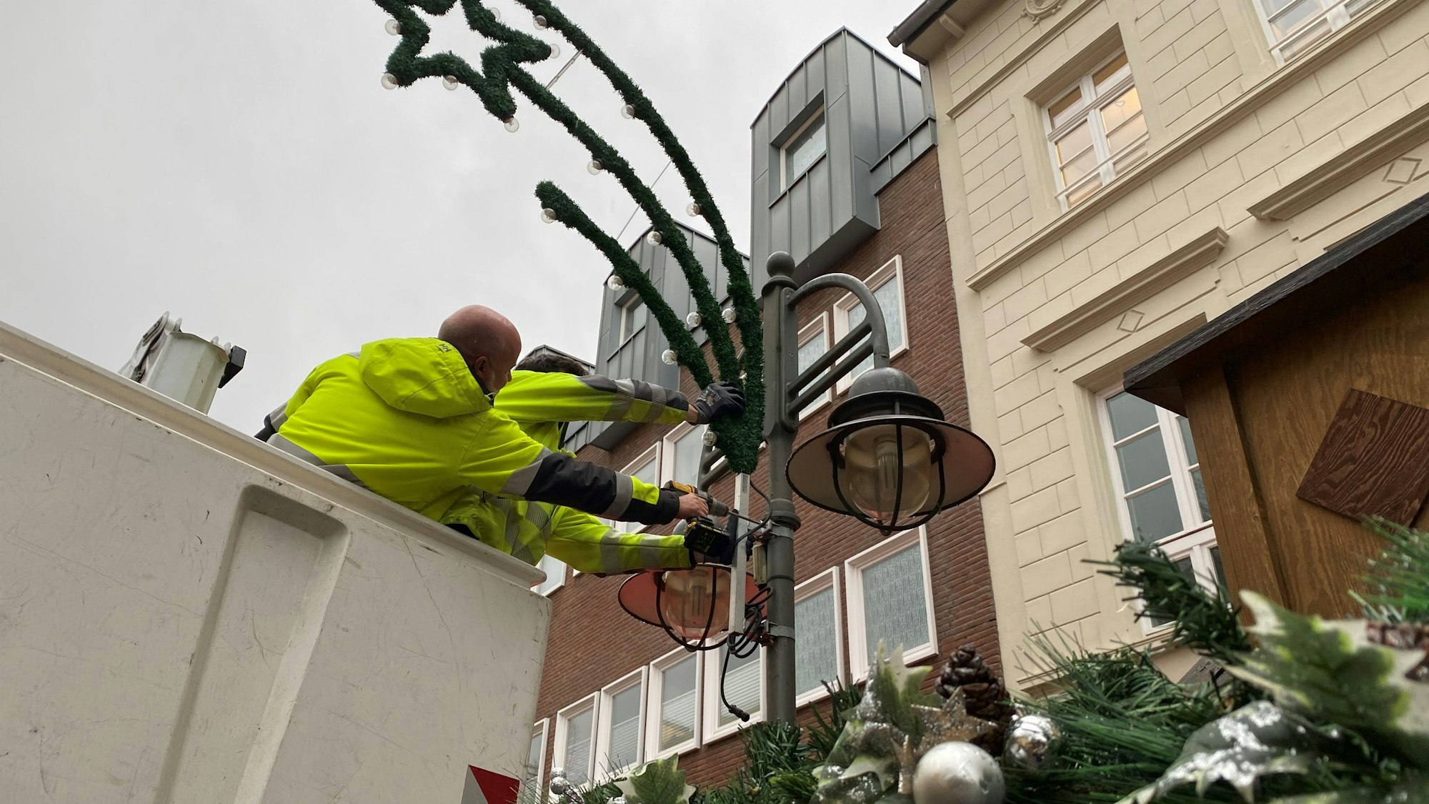 Zu sehen ist, wie zwei Stadtwerke-Mitarbeiter die Weihnachtsbeleuchtung an einem Laternenmast anbringen.