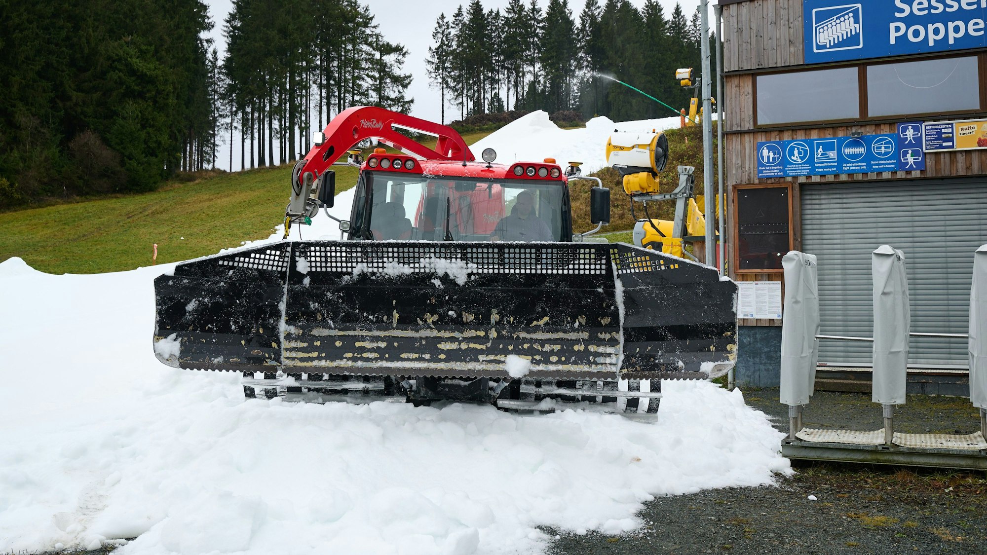 Vor einer Liftanlage steht eine Pistenraupe auf einem Schneeberg. Dahinter ist eine grüne Wiese zu sehen.