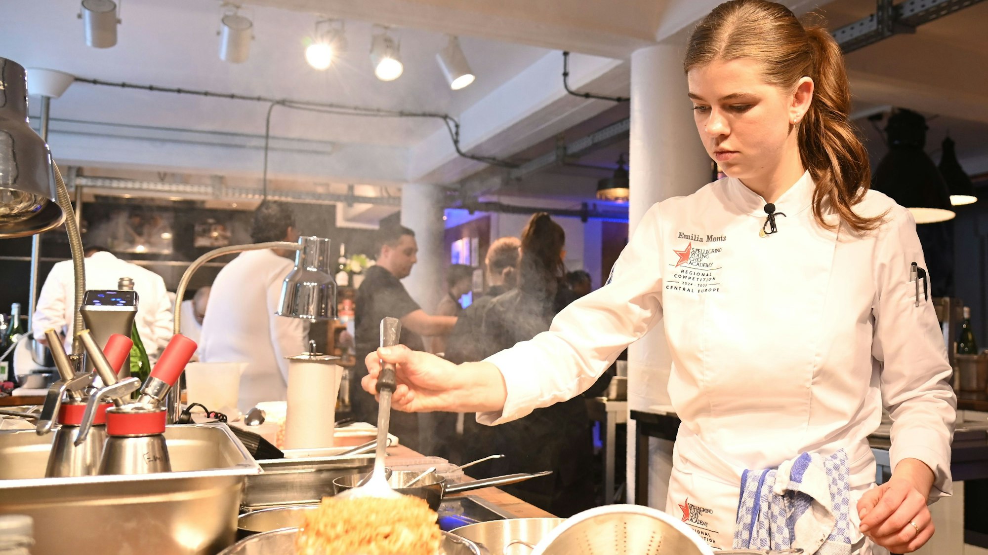 Eine junge Frau in weißer Kochjacke steht vor einem Herd.