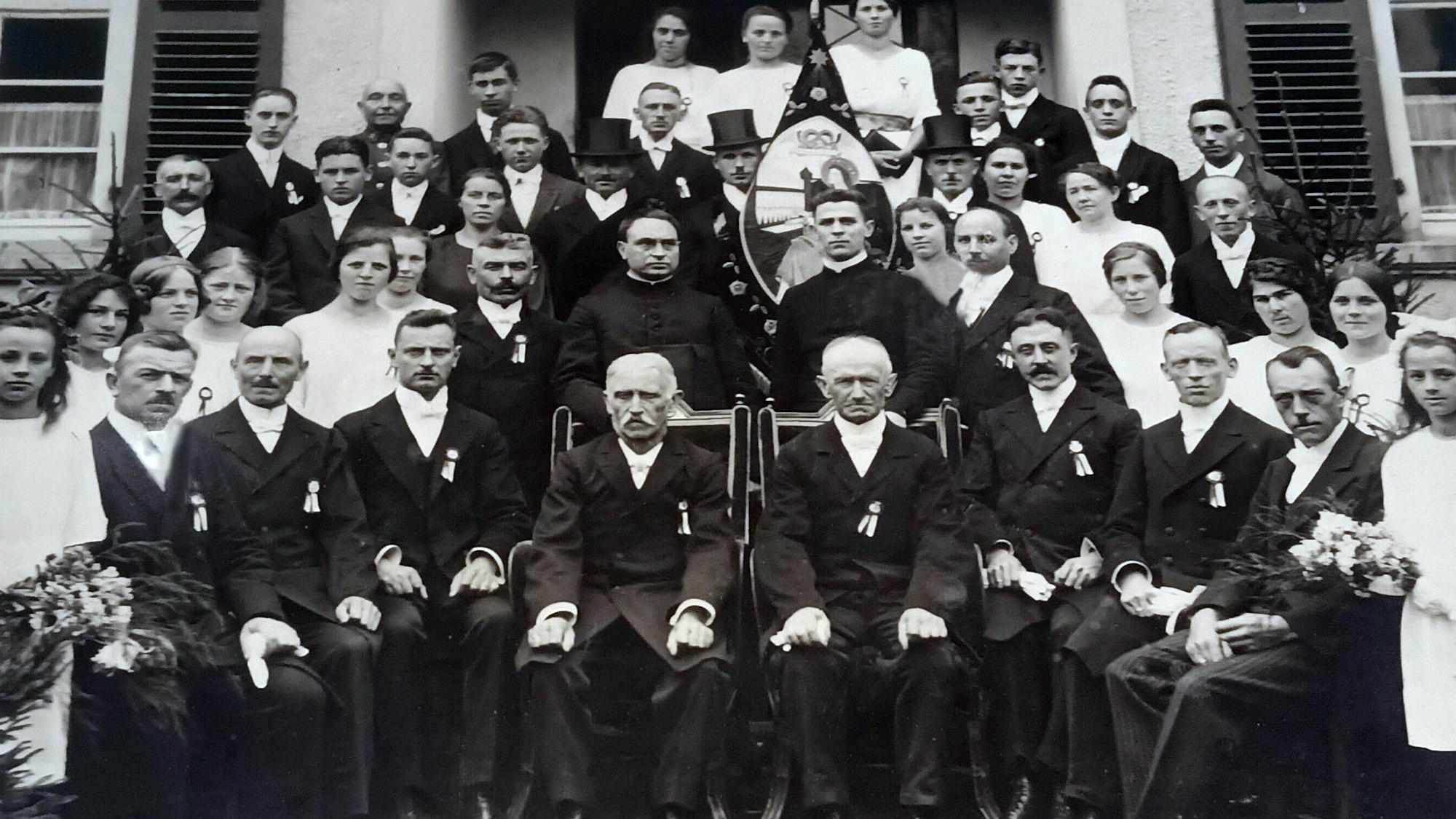 Auf einer Schwarz-Weiß-Aufnahme sitzen und stehen Männer in dunklen Anzügen und Mädchen und Frauen in weißen Kleidern vor einem Gebäude.