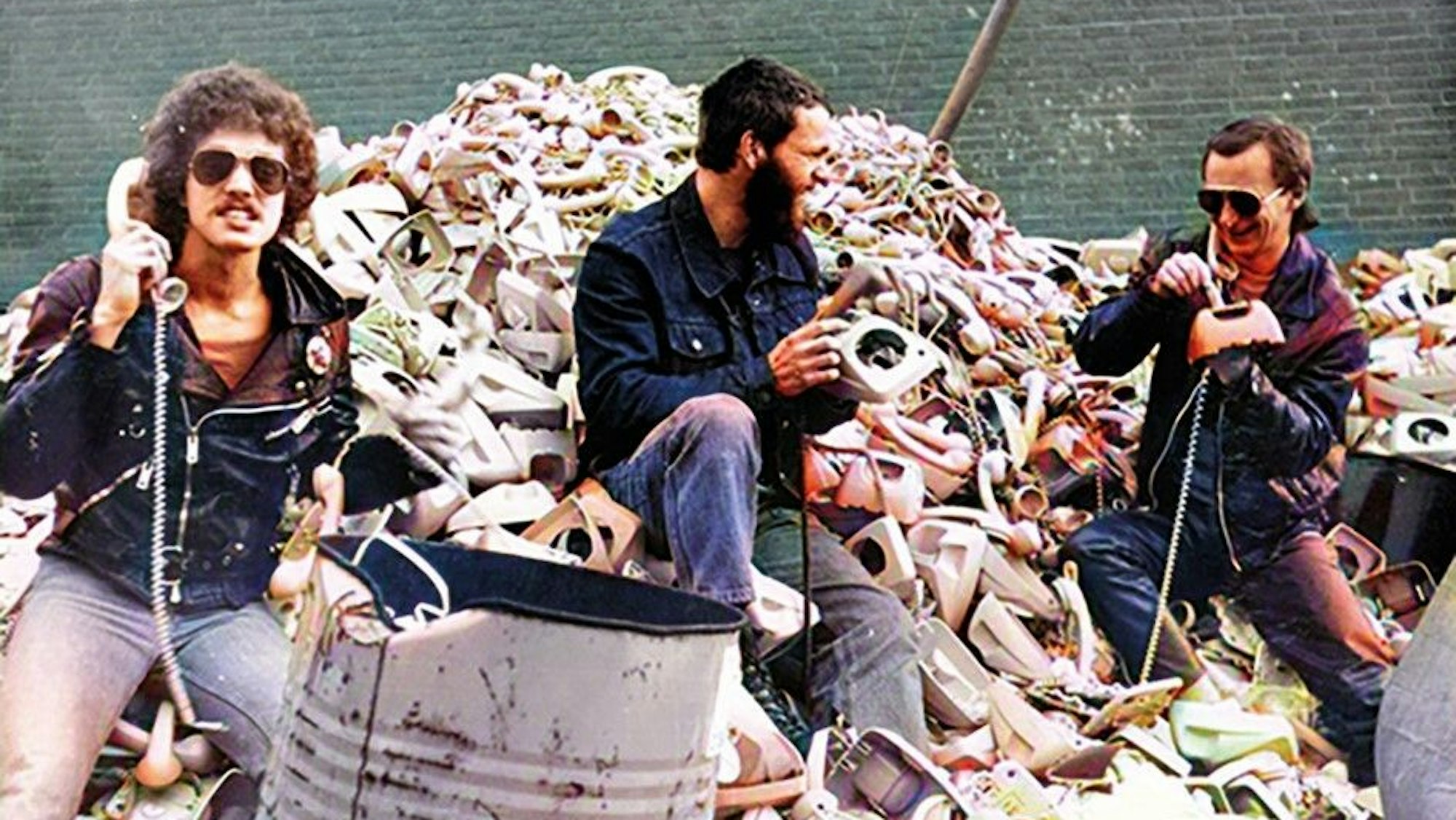 Drei Musiker sitzen mit Telefonen auf einem Müllberg mit Telefonen.