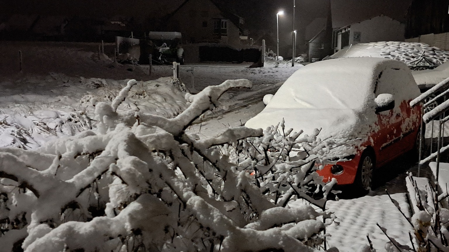 Ein Baum, ein Auto und eine Straße sind von einer Schneedecke überzogen. Es ist Nacht und eine Straßenlaterne leuchtet.