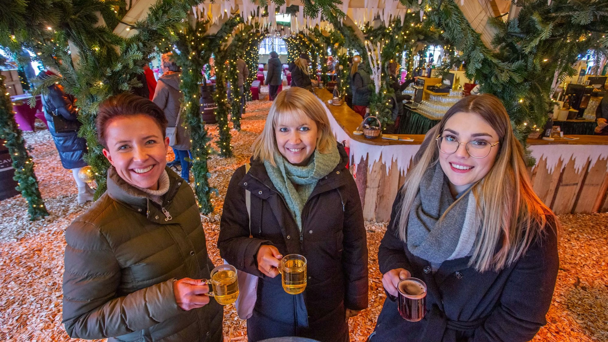 Drei Frauen stehen in einer Weihnachtshütte beisammen. Alle haben einen Glühwein in der Hand.