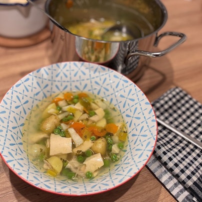 Hühnersuppe in einer Schale, dahinter steht ein Topf.