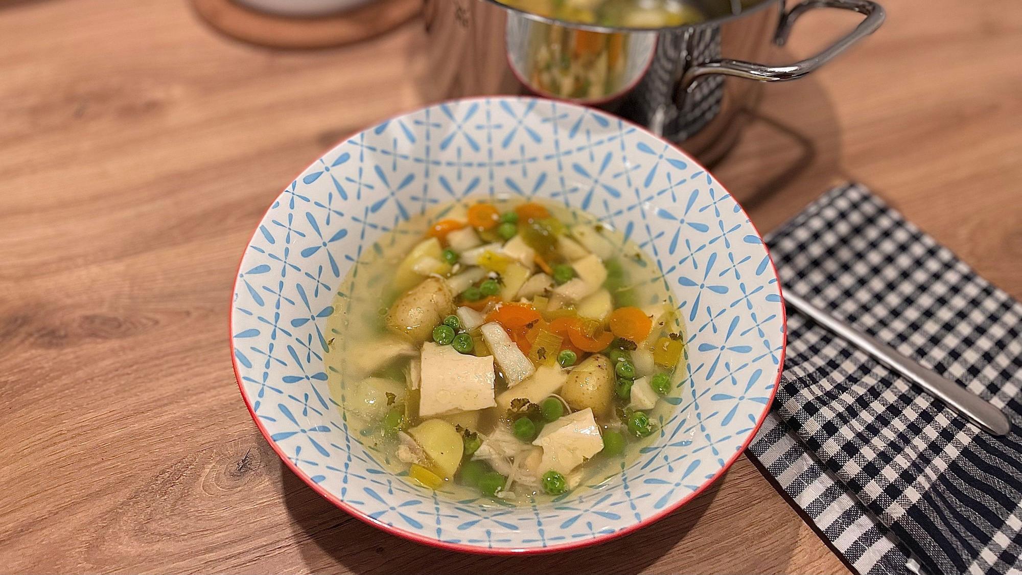 Hühnersuppe in einer Schale, dahinter steht ein Topf.