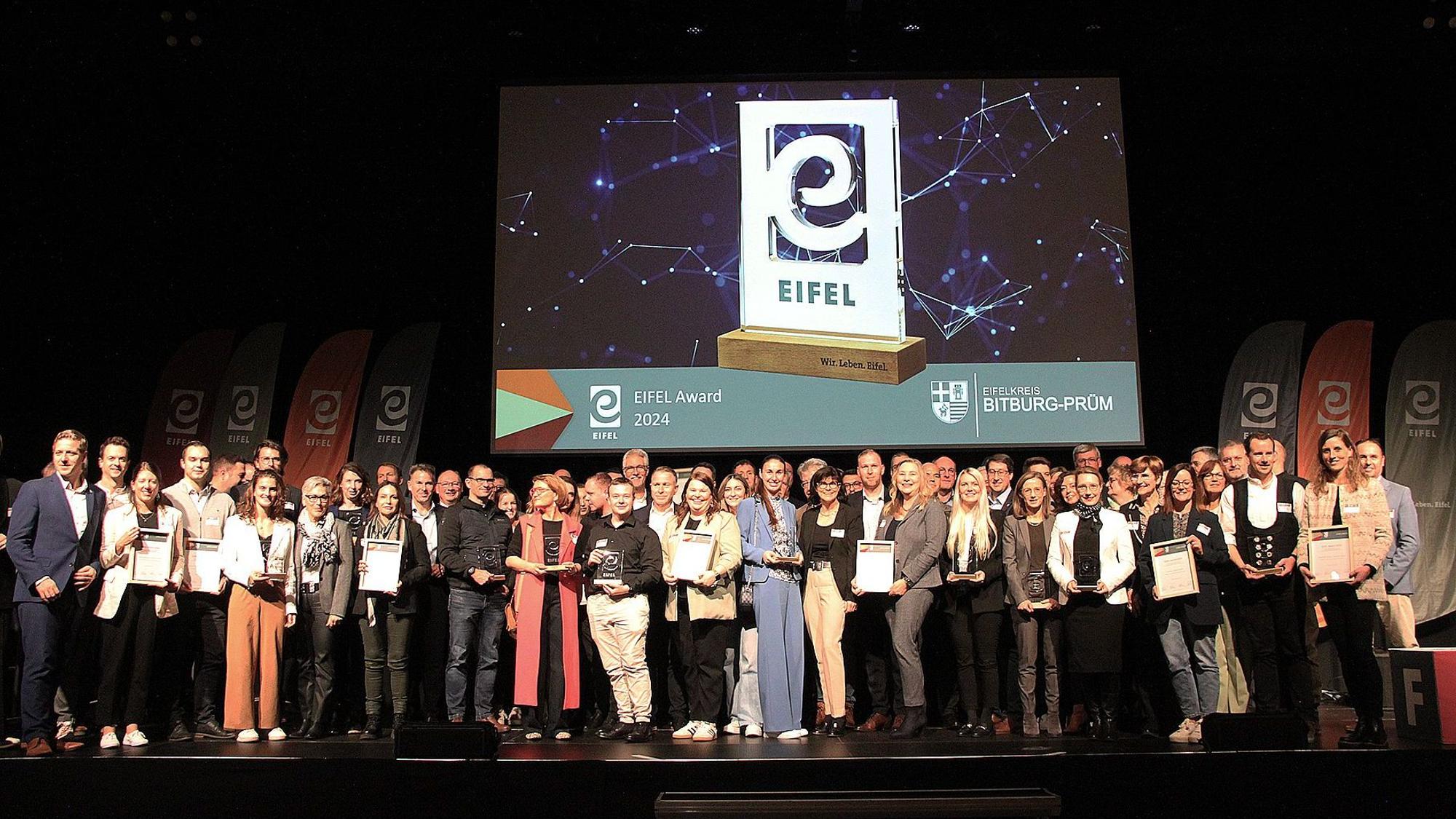 Auf einer Bühne stehen viele Menschen dicht bei andern. Im Hintergrund zeigt ein Bildschirm den Preis der Eifel Awards.
