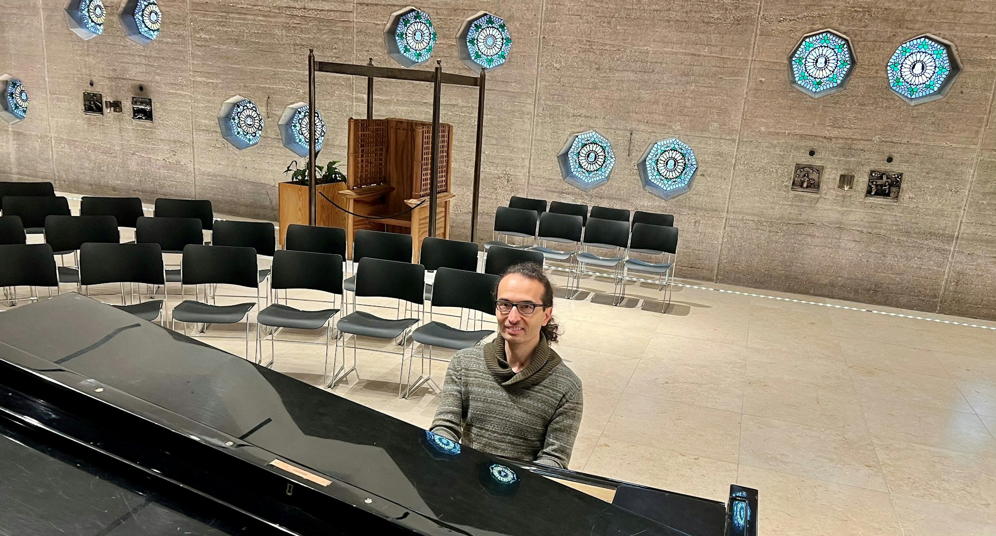Ein Mann mit Zopf und Brille sitzt an einem Flügel in einem runden Raum, der zahlreiche achteckige Fenster hat.