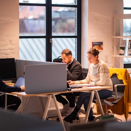 Das Bild zeigt drei Menschen an Schreibtischen. Sie arbeiten ihren Laptops.