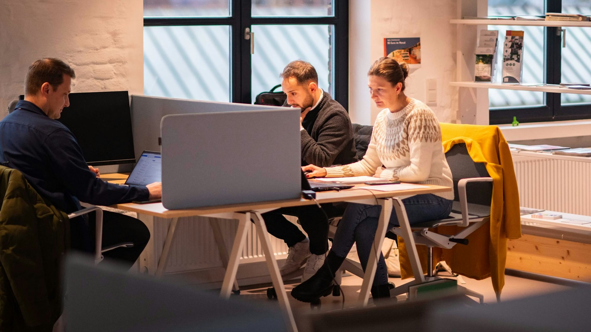 Das Bild zeigt drei Menschen an Schreibtischen. Sie arbeiten ihren Laptops.