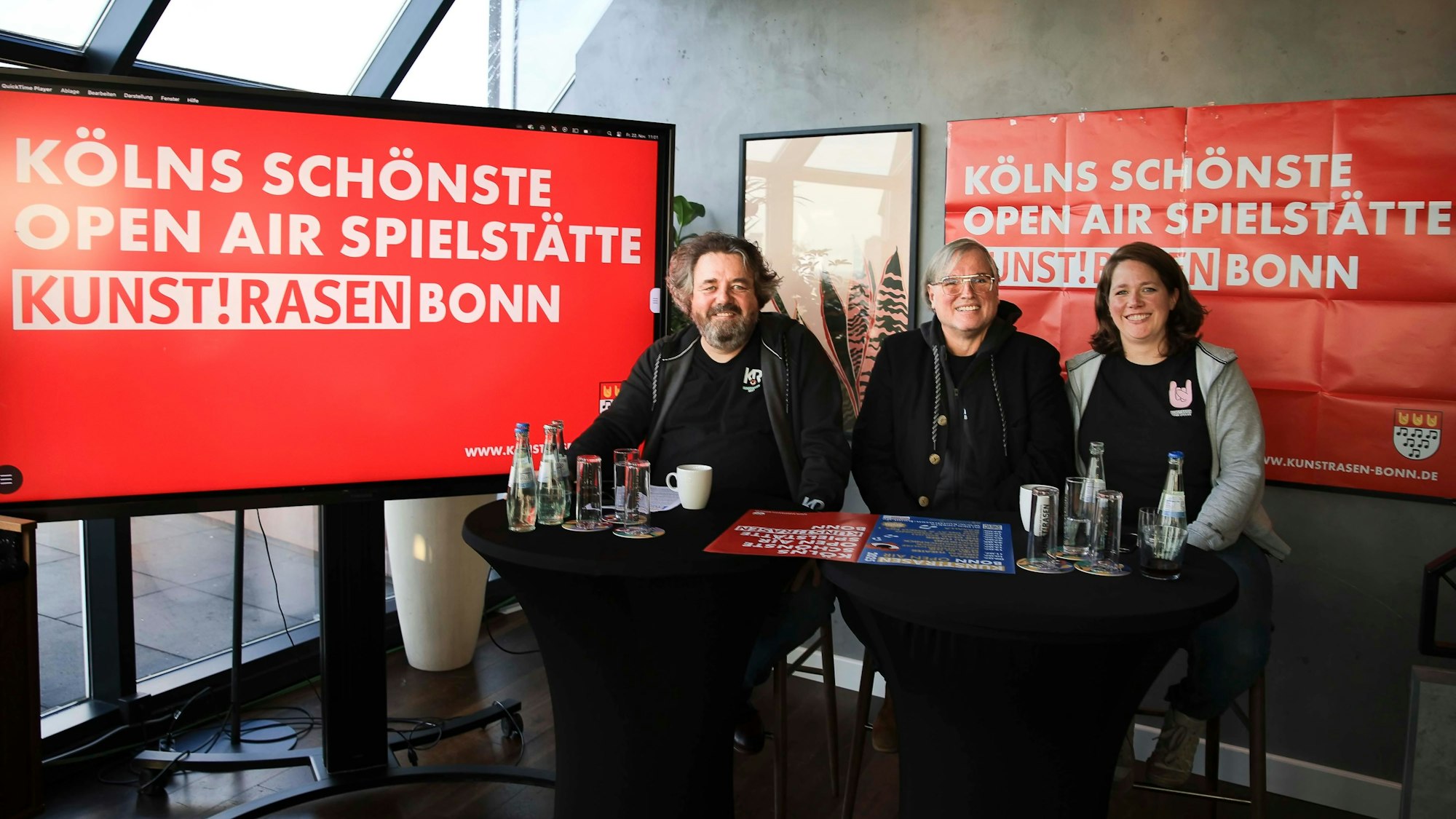Von links nach rechts: Martin J. Nötzel, Ernst-Ludwig Hartz und Katrin Wurm sitzen vor einem Bildschirm. Dort steht: „Kölns schönste Open Air Spielstätte. Kunstrasen Bonn.“