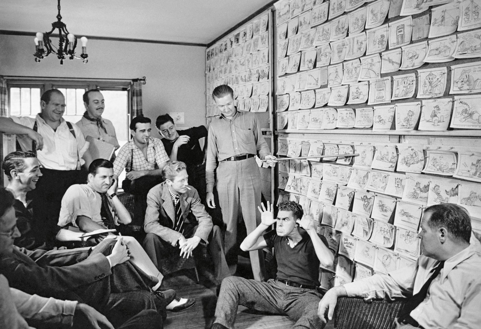 Trickfilm-Zeichner der Walt-Disney-Studios sitzen vor einer Wand mit Einzelzeichnungen. Zeichner Harry Reeves sitzt davor auf dem Boden und zieht eine Grimasse. Carl Barks steht und verweist mit dem Zeigestab auf eine Zeichnung.
