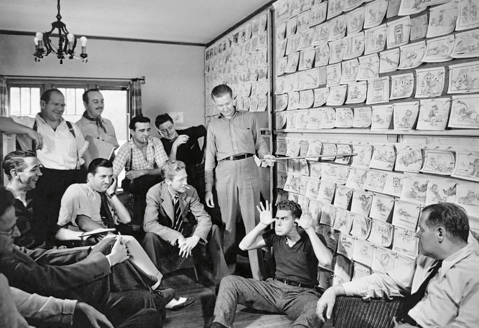 Trickfilm-Zeichner der Walt-Disney-Studios sitzen vor einer Wand mit Einzelzeichnungen. Zeichner Harry Reeves sitzt davor auf dem Boden und zieht eine Grimasse. Carl Barks steht und verweist mit dem Zeigestab auf eine Zeichnung.