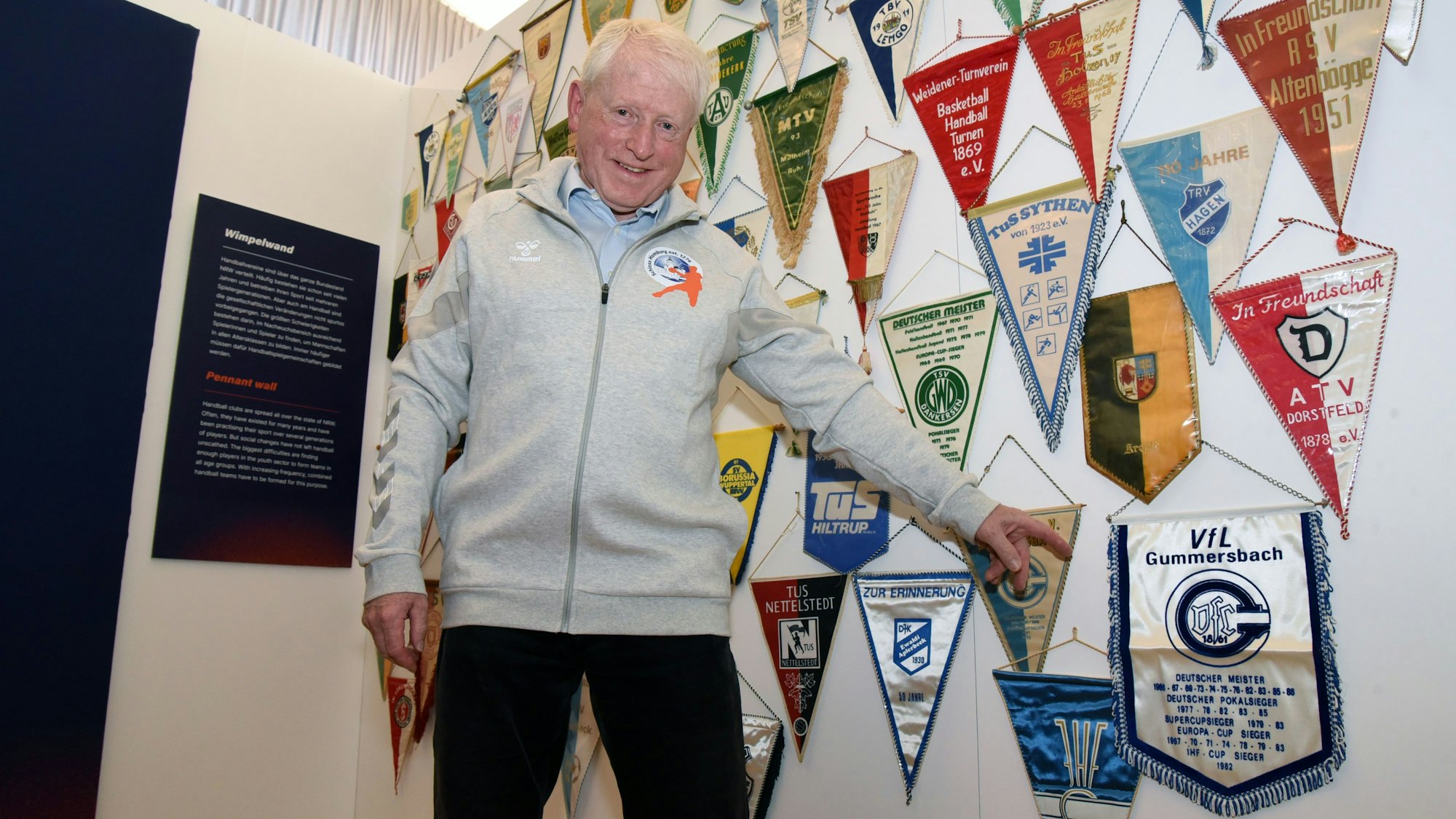 Karl Höver steht vor einer Sammlung zahlreicher Handball-Wimpel und zeigt auf eines des VfL Gummersbach.