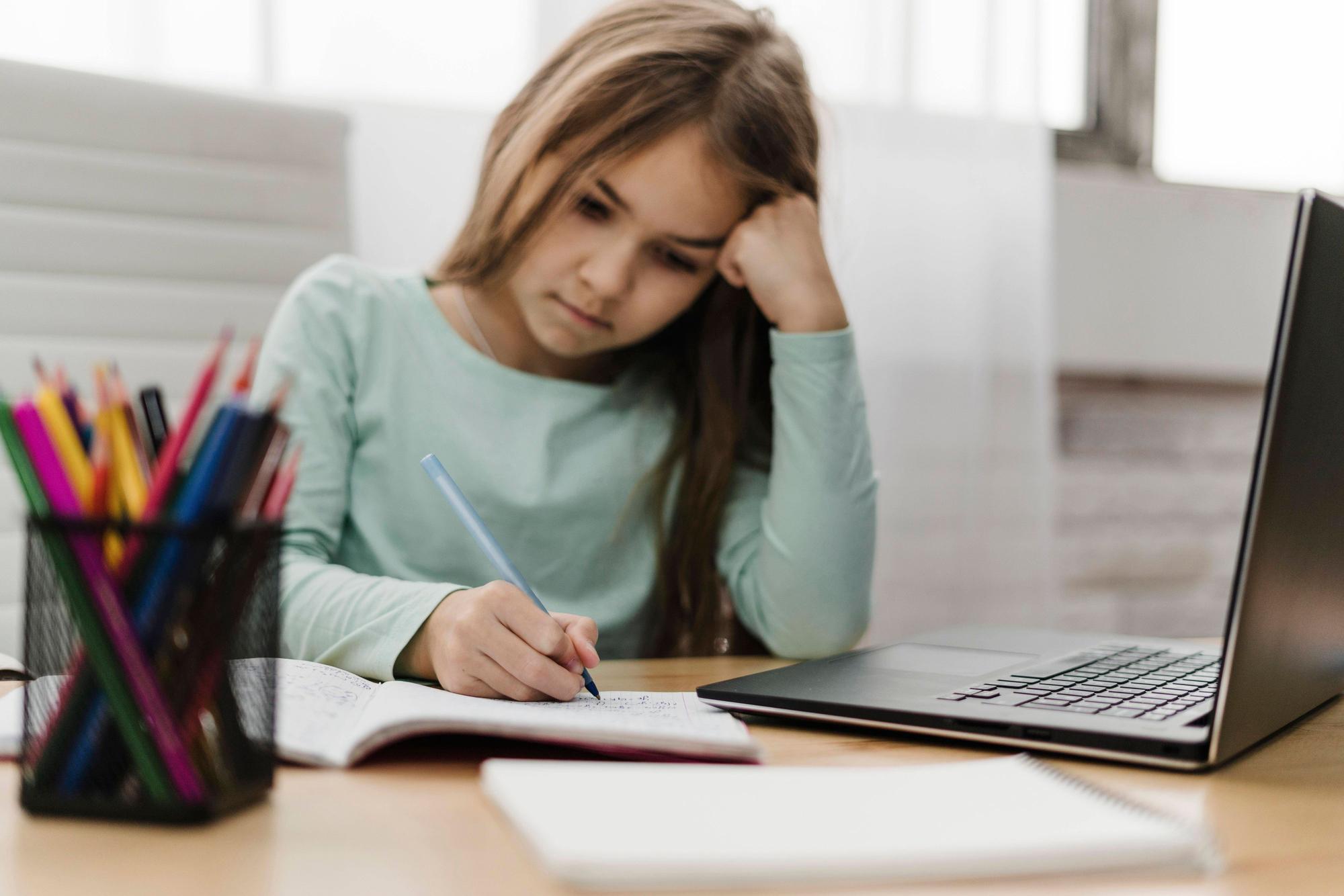 Ein Mädchen sitzt an einem Schreibtisch, auf dem ein Laptop und eine Stifte-Box stehen. Das Mädchen notiert etwas in ein Heft; es nimmt offenbar an einem Online-Unterricht teil.