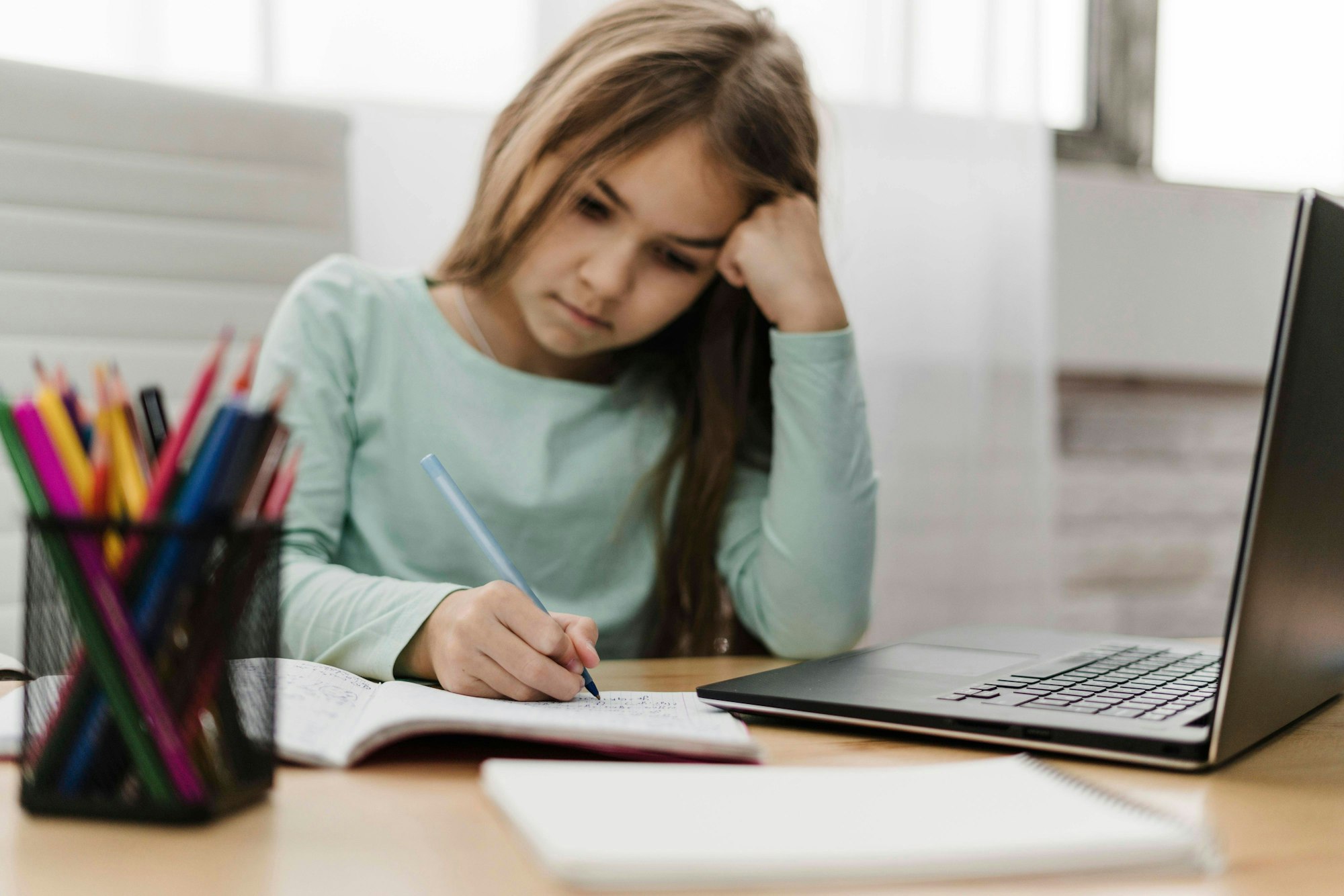 Ein Mädchen sitzt an einem Schreibtisch, auf dem ein Laptop und eine Stifte-Box stehen. Das Mädchen notiert etwas in ein Heft; es nimmt offenbar an einem Online-Unterricht teil.