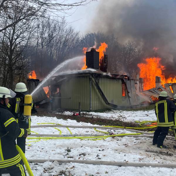 Feuerwehrleute löschen eines der langgestreckten Gebäude in der ZUE in Vogelsang. Die Flammen lodern aus dem Dach.