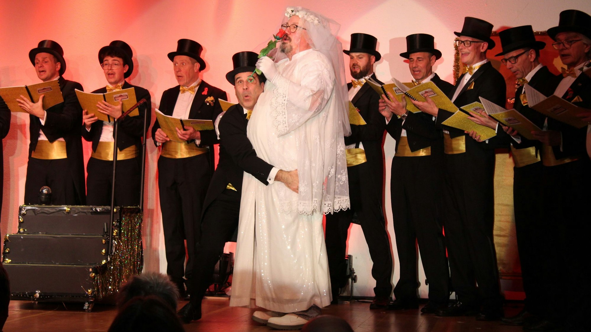 Auf dem Bild sind ein als Braut verkleideter Mann und viele Chorsänger im Frack zu sehen.