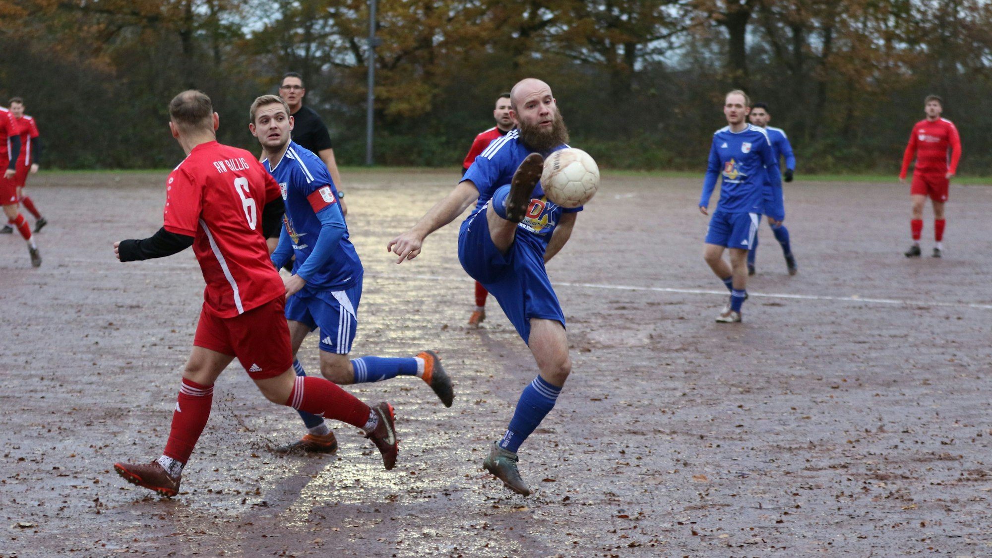 Ein Spieler passt den Ball mit einem hohen Bein, das waagerecht in der Luft steht. Der Aschenplatz ist komplett nass.
