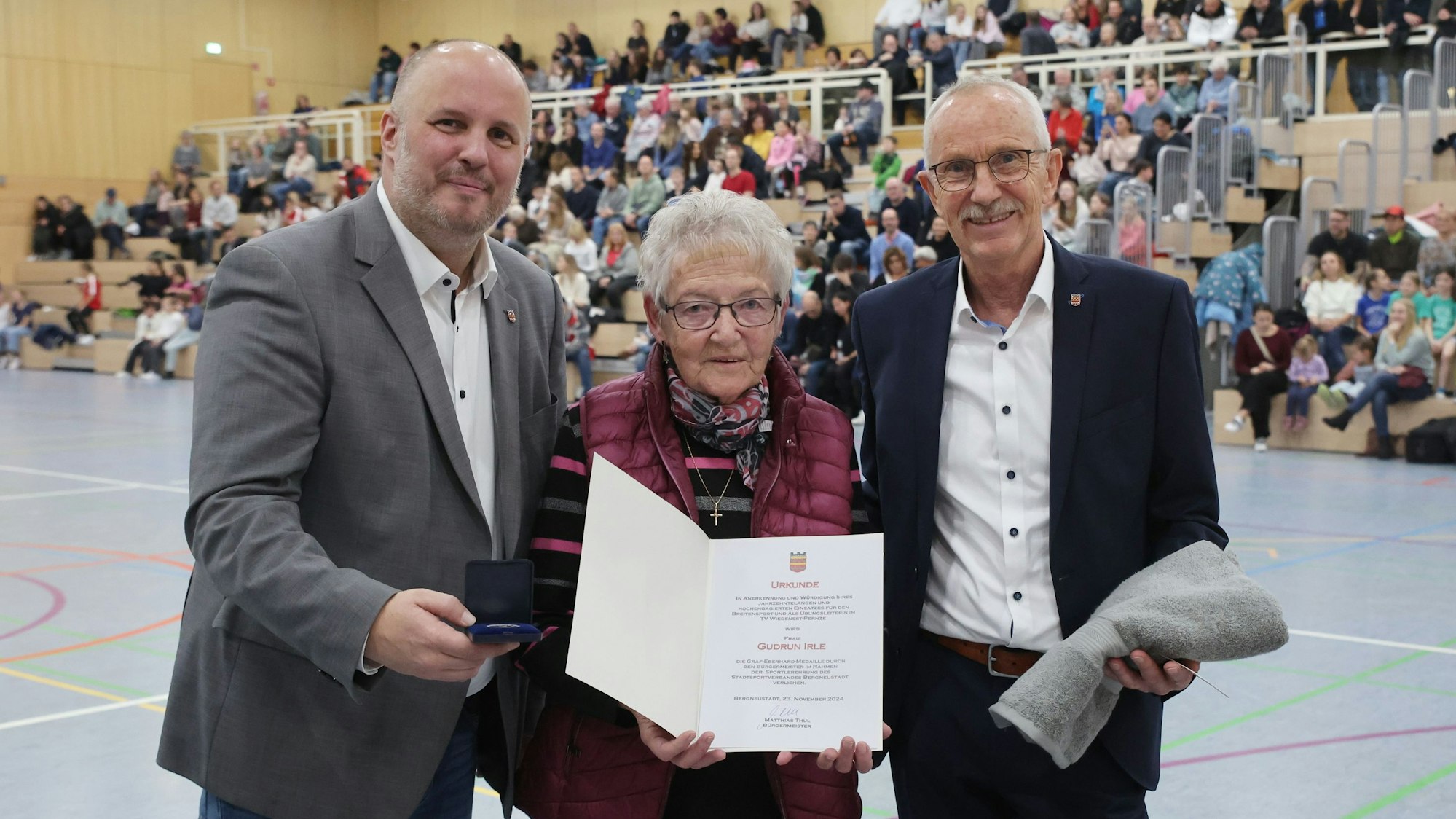 Zwei Herren und eine Dame zeigen Urkunde und Medaille.