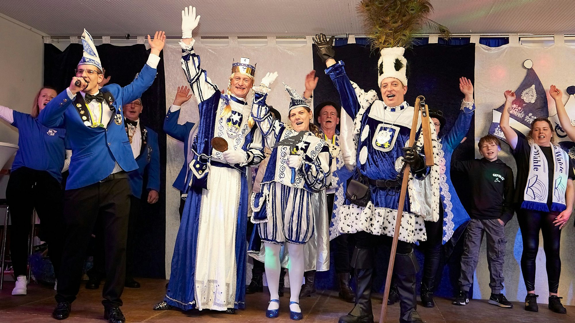 Das Wahlener Dreigestirn mit Prinz Petra I, Bauer Daniel und Jungfrau Erika steht zusammen mit weiteren Karnevalisten im blau-weißen Ornat auf der Bühne und winkt ins Publikum.