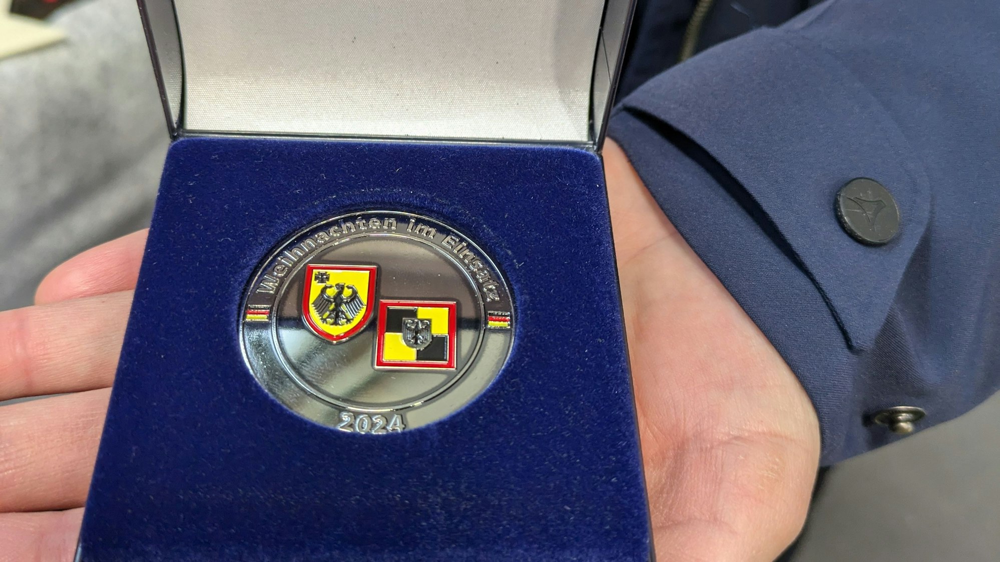 Eine Hand hält die Schatulle mit dem Einsatz-Coin ins Bild. Auf dem Coin befinden sich zwei Wappen und die Aufschrift „Weihnachten im Einsatz 2024“.
