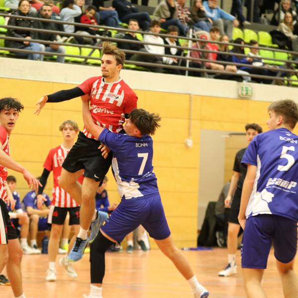 Der geworfene Ball eines Handballers, der in der Luft steht, landet im Gesicht des Gegenspielers. Ein weiterer Gegenspieler hat sich beim Werfer eingehakt.