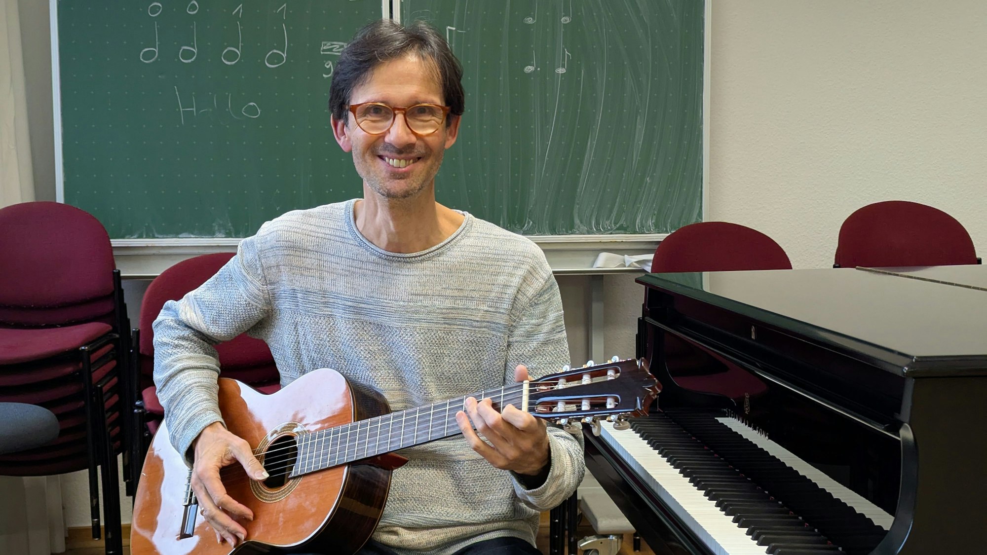 Das Foto zeigt einen Mann mit Gitarre neben einem Piano, im Hintergrund eine Tafel mit Noten.