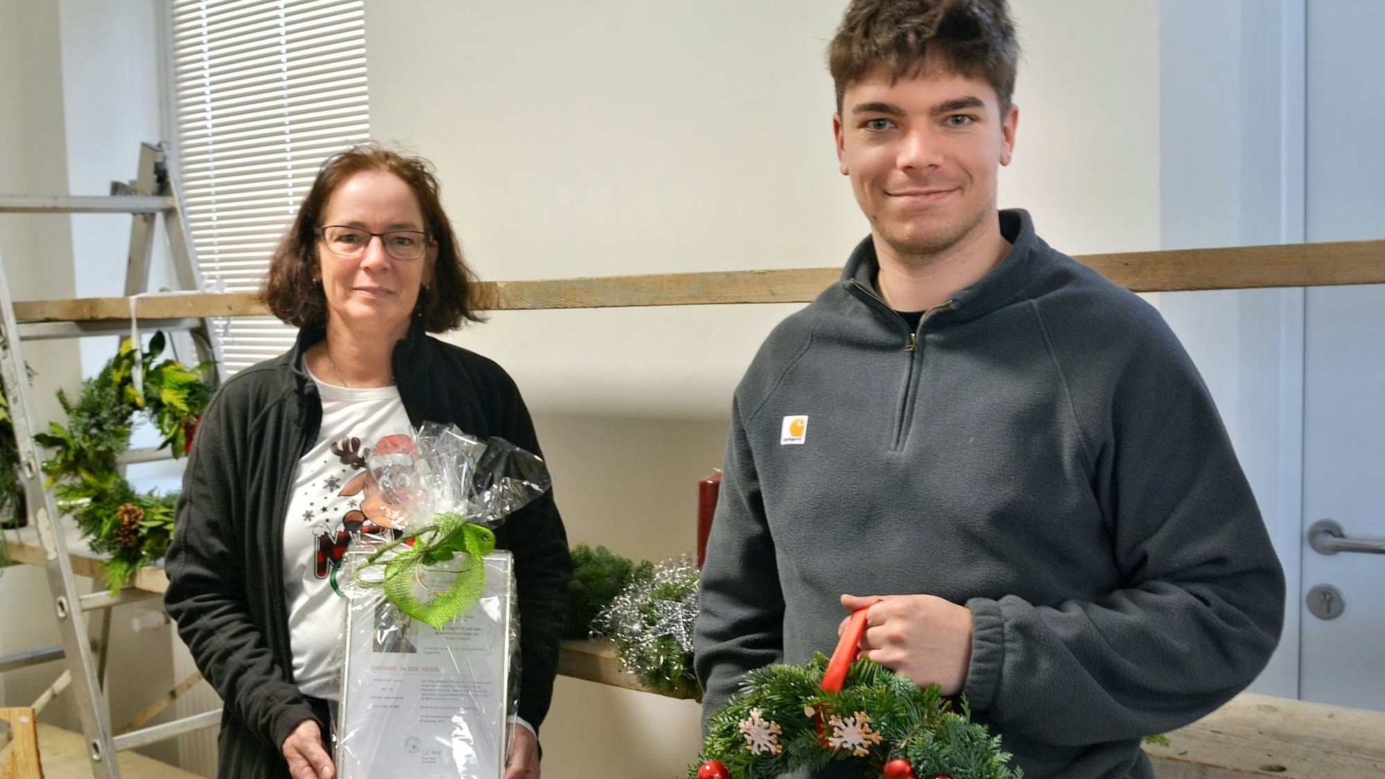 Michaela Lingscheid zeigt eine in Folie verpackte, gerahmte Urkunde, ihr Sohn Marko einen aus Tannengrün geflochtenen Kranz.