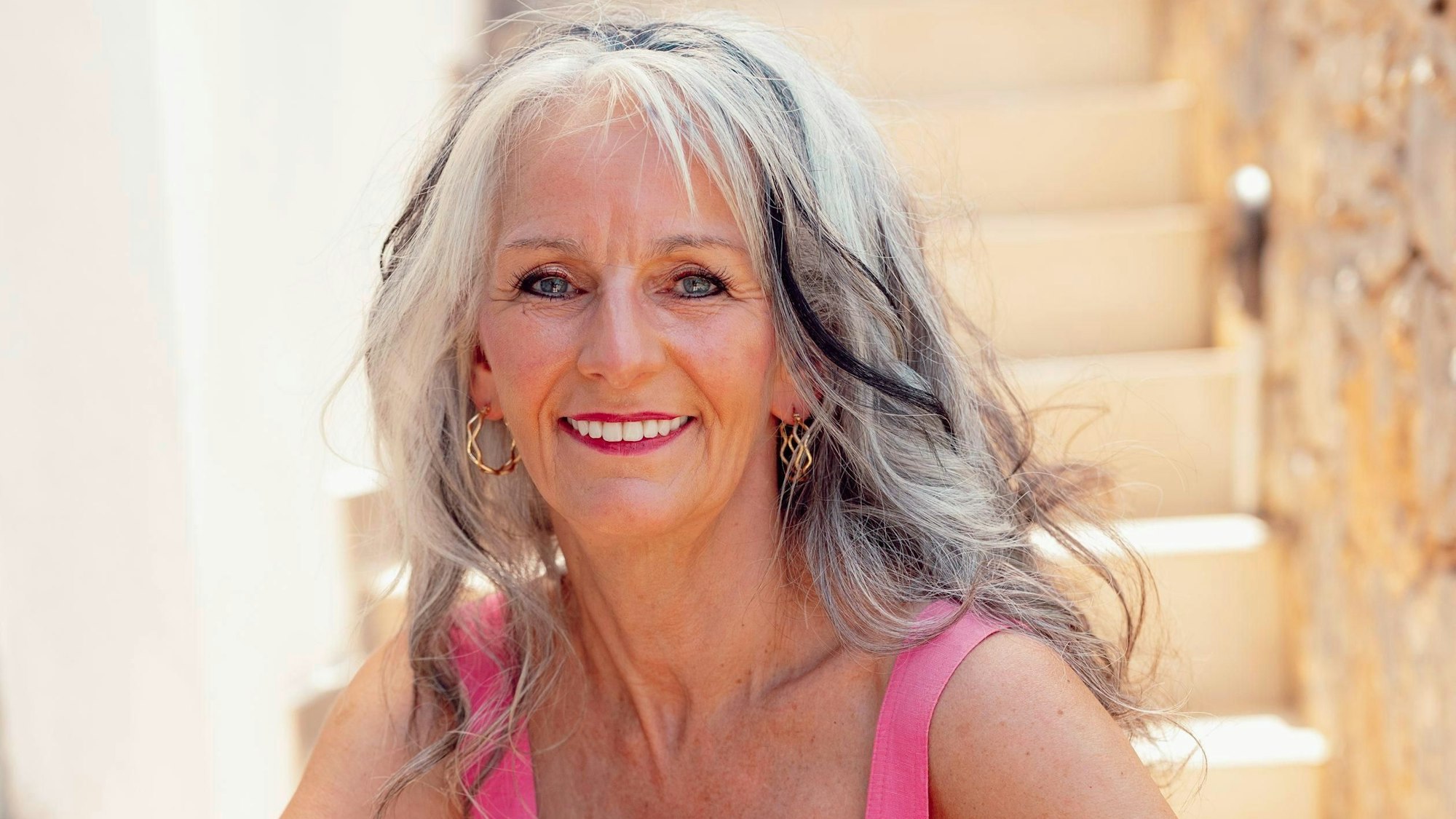 Sabine aus Troisdorf ist dreifache Mutter und eine von 18 Kandidatinnen der RTL-Datingshow „Golden Bachelor“. Sie schaut auf dieser Fotografie lächelnd in die Kamera, sitzt dabei auf einer Treppe.