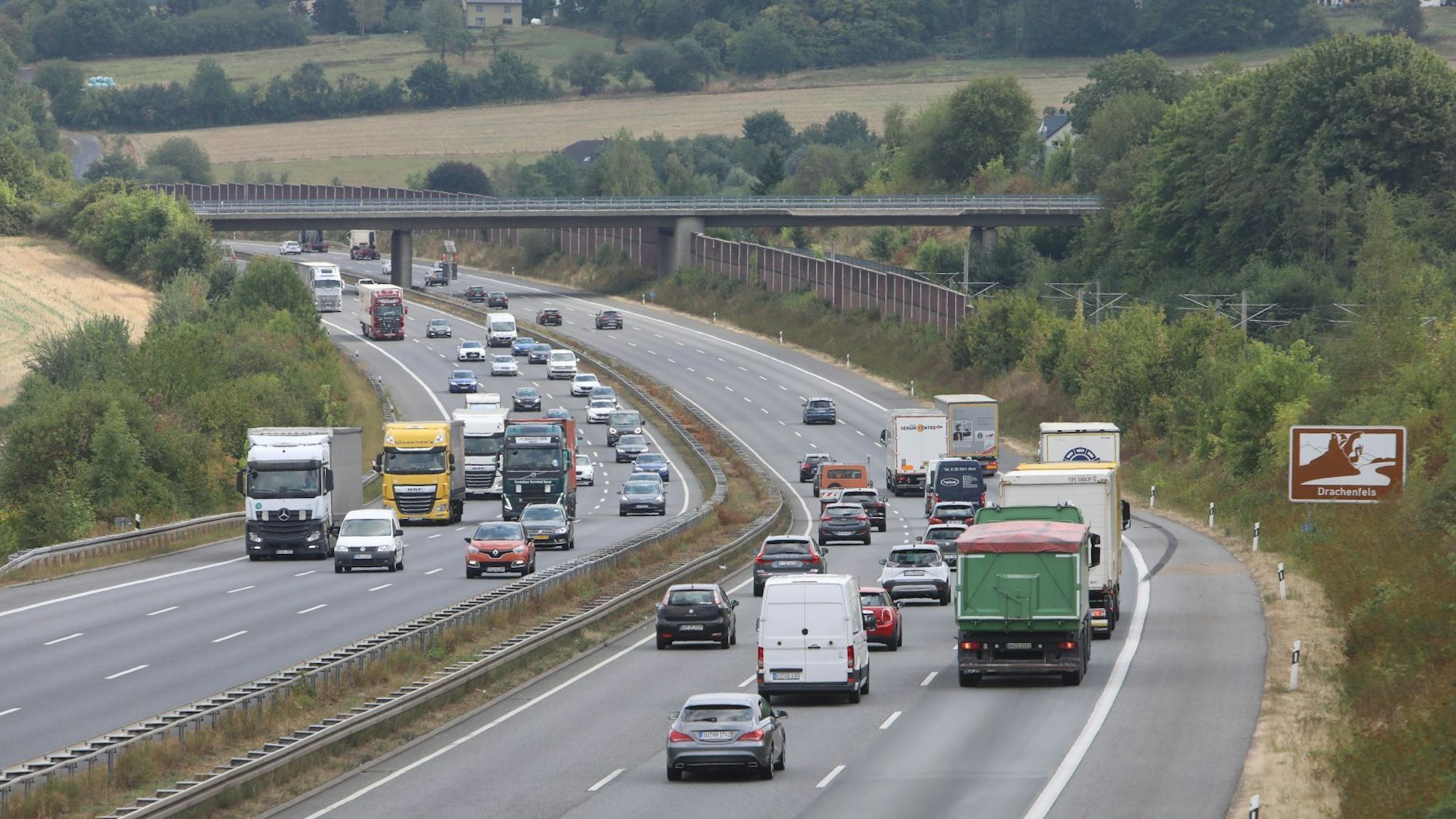 Pkw und Lkw auf einer sechsspurigen Autobahn, ein Schild am Fahrbahnrand weist auf den Drachenfels hin.