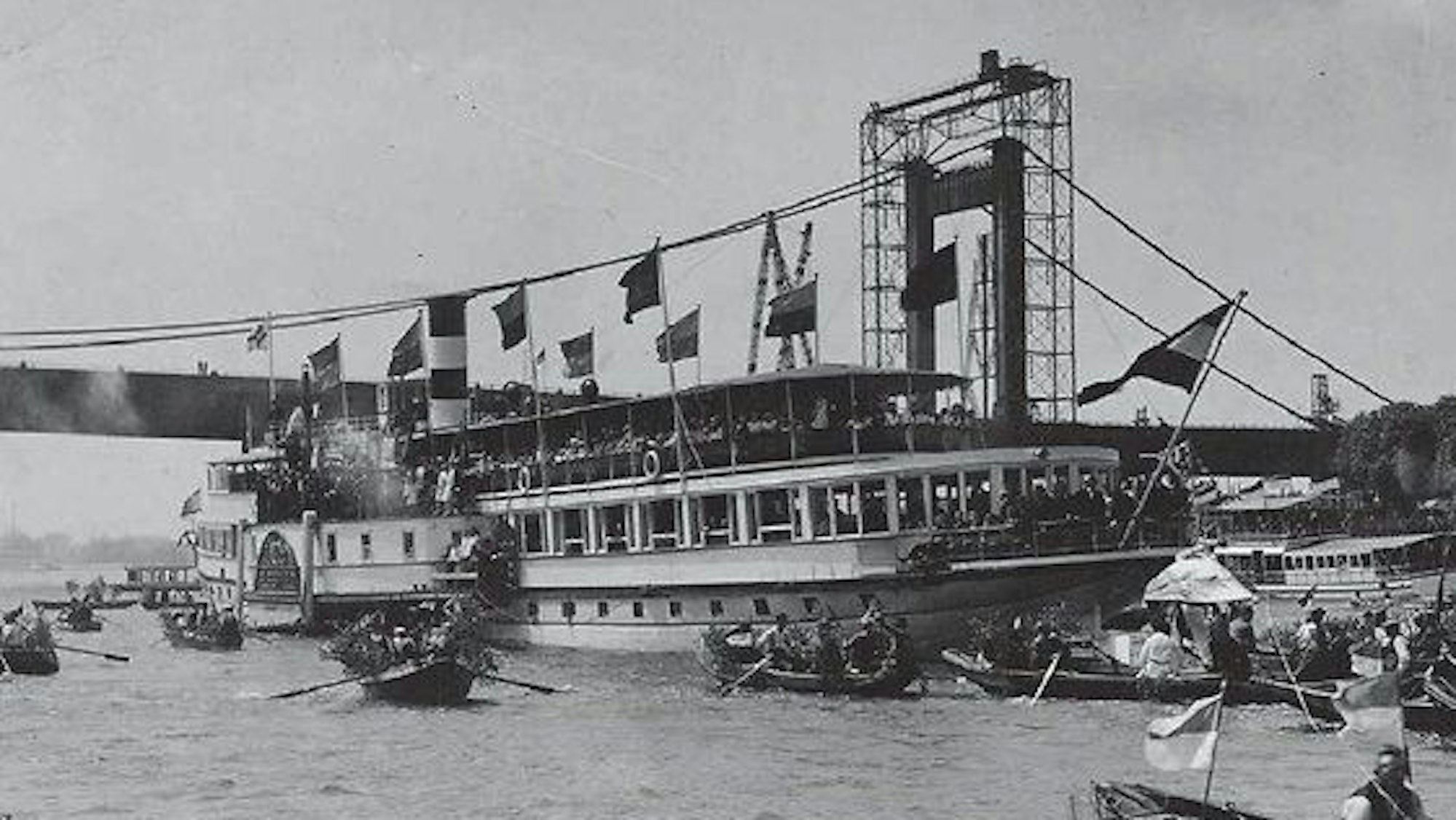 Ein Schwarz-weiß-Bild zeigt ein großes fahnengeschmücktes Schiff begleitet von vielen kleinen Booten.